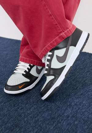 Osoba nosząca czarno-białe buty sportowe z pomarańczowym logo Nike i czerwone spodnie stojąca na niebieskim dywanie.