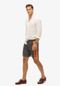 Witte linnen overhemd, grijze korte broek met oranje accent, met de opdruk 'SUPERDRY SPORTSWEAR'. Bruine bootschoenen maken de look compleet.