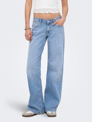 Person som har på seg lyseblå vide jeans, hvit blonder crop topp og grå-hvite joggesko, står med en hånd i lomma.