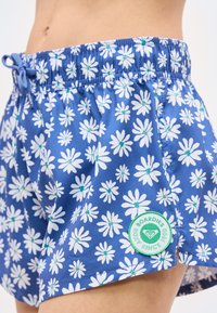 Μπλε σορτς με λευκά σχέδια μαργαρίτας και πράσινο κυκλικό λογότυπο της μάρκας Roxy, φορεμένα από άτομο με ορατή κοιλιά.