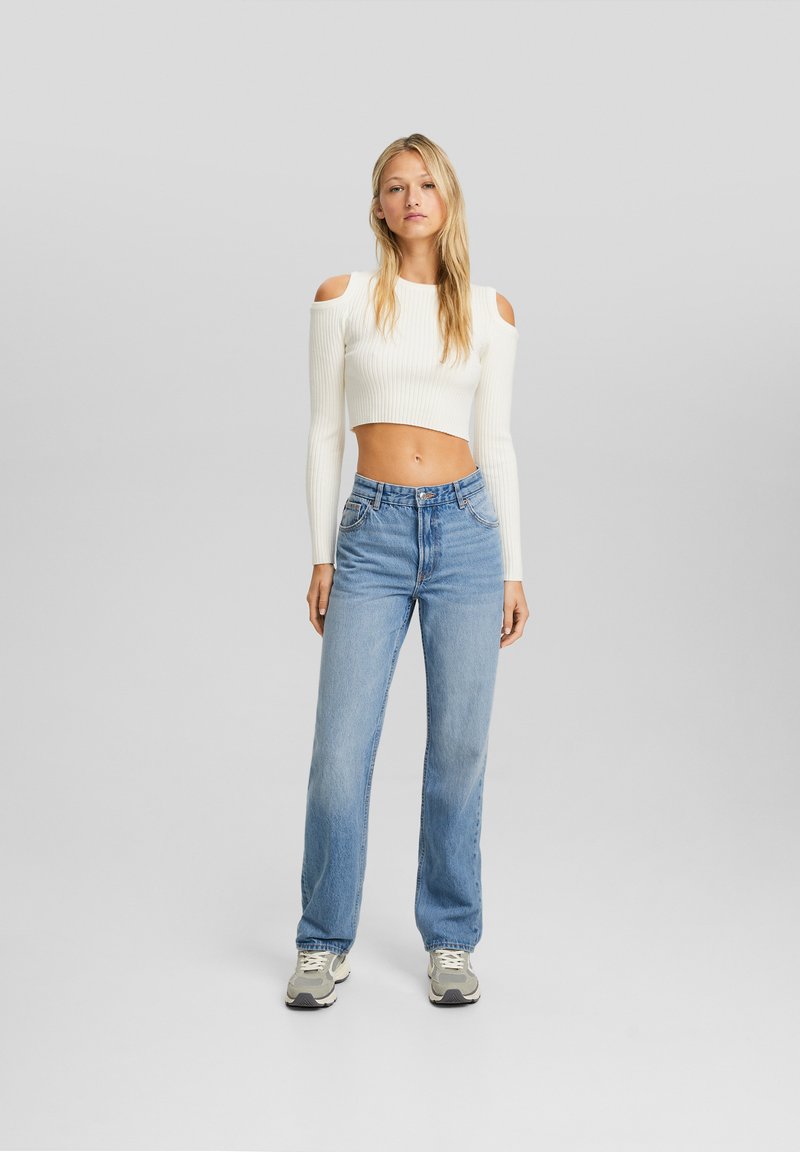 Biały prążkowany crop top z odkrytymi ramionami, w połączeniu z jasnoniebieskimi jeansami o prostym kroju. Szare trampki dopełniają stylizację.