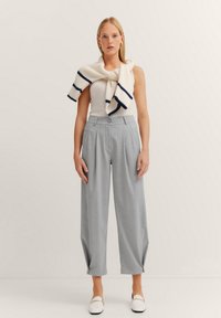 Helle graue, gefaltete, hoch-taillierte Hose kombiniert mit einem weißen ärmellosen Oberteil und einem cremefarbenen Strickpullover mit marineblauen Streifen, der über die Schultern drapiert ist.