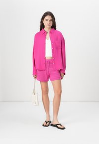 Camicia oversize rosa brillante con bottoni, canotta bianca, pantaloni corti rosa a trama, infradito nere e una borsa bianca. Tessuto morbido, stile casual.
