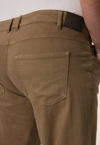I pantaloni in denim marroni presentano un design aderente, due tasche posteriori con cuciture curve e un'etichetta in pelle scura sulla cintura.