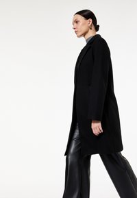 Abrigo de lana negro con un diseño ajustado, que presenta mangas largas y una silueta recta. Usado sobre pantalones de cuero negros, de estilo minimalista.