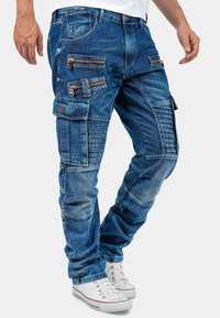 Pantalons cargo en denim bleu avec plusieurs poches zippées, des pièces texturées et une coupe fuselée. Comprend des détails de couture et un aspect légèrement usé.