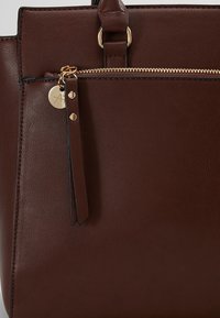 Sac à main en cuir marron avec une tirette de fermeture éclair dorée et un petit pendentif rond en or accroché à la languette de la fermeture éclair.