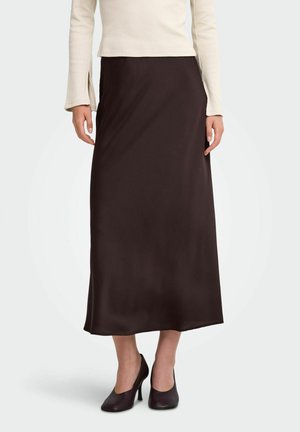 Femme portant une longue jupe en satin marron foncé, un haut crème à manches longues et des chaussures à talons hauts marron foncé, debout sur un fond clair uni.