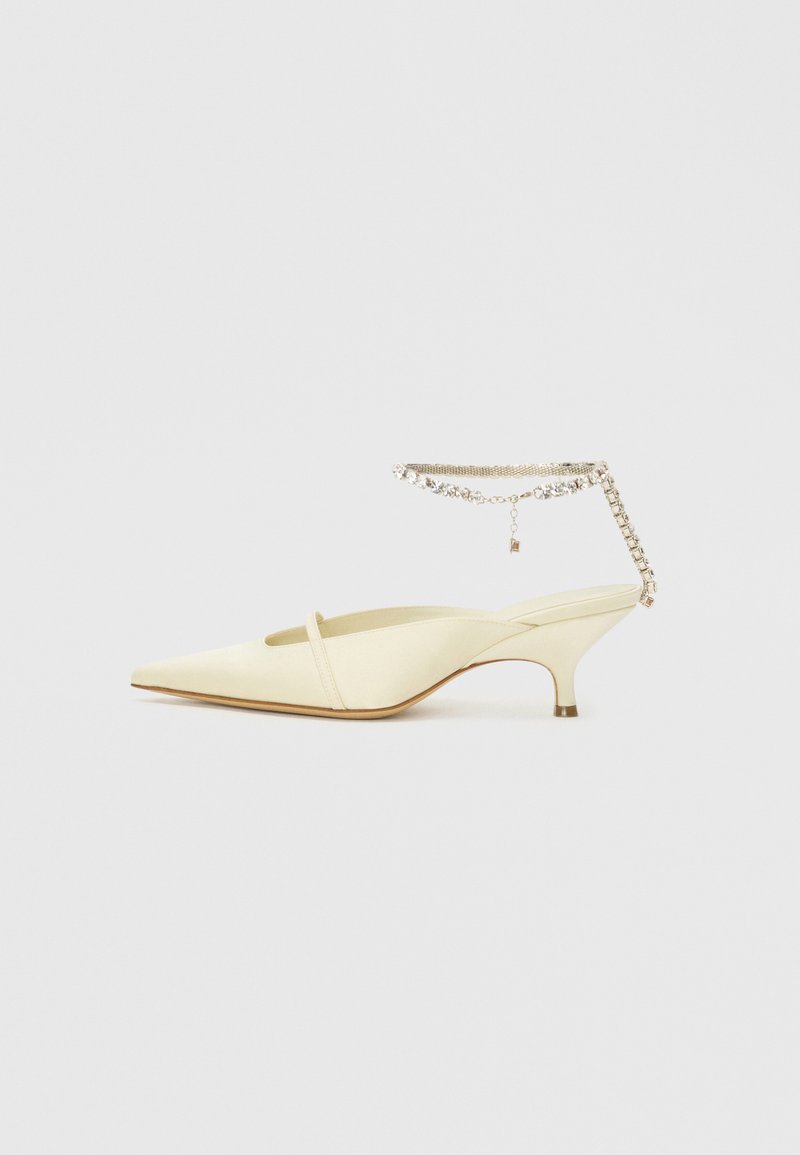 Zapato slingback de satén amarillo con punta afilada, tacón bajo y una correa de tobillo decorativa con cadena que presenta rhinestones. Textura suave, diseño minimalista.