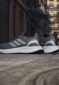 Ζευγάρι γκρι και μαύρα αθλητικά παπούτσια Adidas με λευκές σόλες σε πλακόστρωτο δρόμο, σημειωμένη αδιαβροχοποίηση, αστικό φόντο.