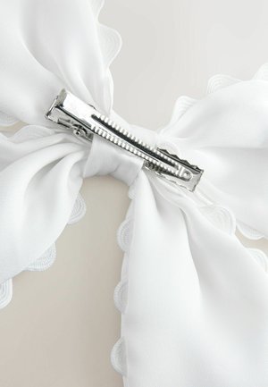White fabric bow with scalloped edges, featuring a metallic silver clip closure. Smooth texture with a soft, flowing design.