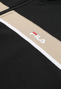 Musta kangas, jossa on beigen ja valkoisen raidat, joista beigen raidassa on valkoinen ja punainen FILA-logo.