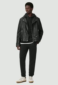 Young man wearing black leather jacket over black hoodie and black joggers, standing with hands in pockets, white sneakers, neutral background.