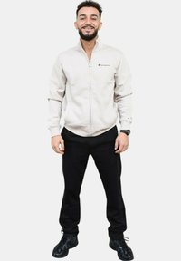 Chaqueta de cremallera gris claro con bolsillos frontales, pantalones negros y zapatillas negras. Presenta un logotipo en el pecho y puños de canalé.