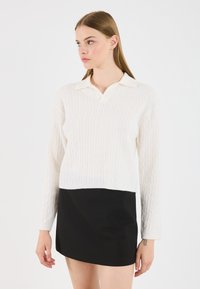 Λευκό πουλόβερ με πλέξη καλωδίου και γιακά, συνδυασμένο με μαύρη μίνι φούστα. Το πουλόβερ έχει μακριά μανίκια και cropped σχεδίαση.