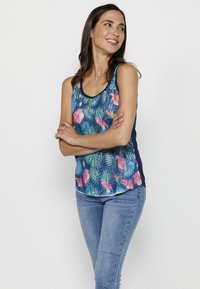 Débardeur fleuri bleu avec des motifs tropicaux roses et verts, présentant un col rond et des panneaux latéraux marine. Associé à un jean en denim clair.