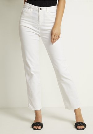 Femme portant un jean blanc droit taille haute et des sandales noires ouvertes à bout tressé, debout sur un fond clair uni.