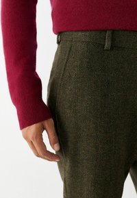 Mörkgrön herringbonebyxor i kombination med en röd stickad tröja. Närbild som visar texturen och detaljerna av tyget och passformen.