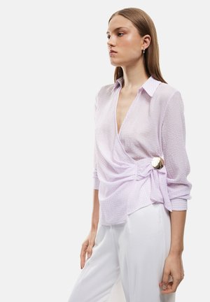 Giovane donna con capelli lisci che indossa una camicetta avvolgente lilla chiaro con un bottone dorato e pantaloni bianchi, in piedi davanti a uno sfondo semplice.