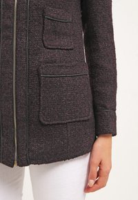 Bunda z vlněné směsi v tmavě fialové barvě s texturovaným povrchem, s předním zipem, černým lemováním a dvěma velkými předními kapsami.