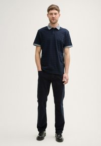 Granatowa koszulka polo z niebiesko-szarym kołnierzykiem i mankietami, z teksturowanymi poziomymi paskami, w połączeniu z ciemnymi spodniami i czarnymi butami, stojąca na jednolitym tle.