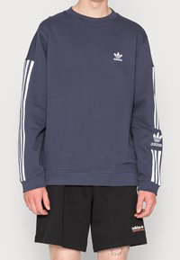 Tummansininen collegepaita, joka on valmistettu puuvillasta. Paidassa on valkoiset kolme raitaa hihoissa ja pieni valkoinen Adidas-logo rinnassa.