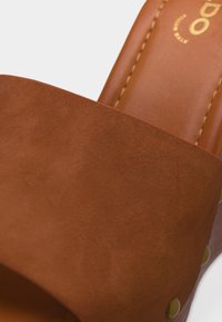 Gros plan d'une sandale à enfiler en cuir marron avec des clous dorés visibles et une semelle intérieure cousue.