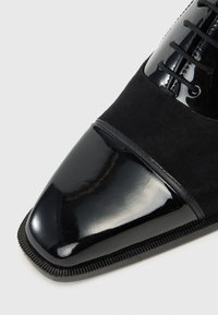 Black dress shoe featuring a sleek patent leather toe, suede upper, and lace-up design. Textured sole with fine stitching detail.