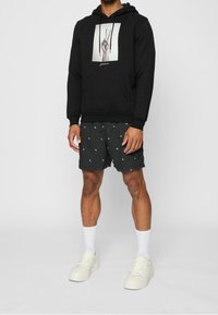 Felpa nera con grafica frontale di una mano che fa il segno della pace, abbinata a pantaloncini grigi con motivo bianco e calzini bianchi.