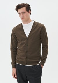 Rjav cardigan z V-izrezom, gumba spredaj in rebrastimi manšetami. Nosen čez belo srajco, ki se ponaša z mehko teksturo in prilegajočim se dizajnom.