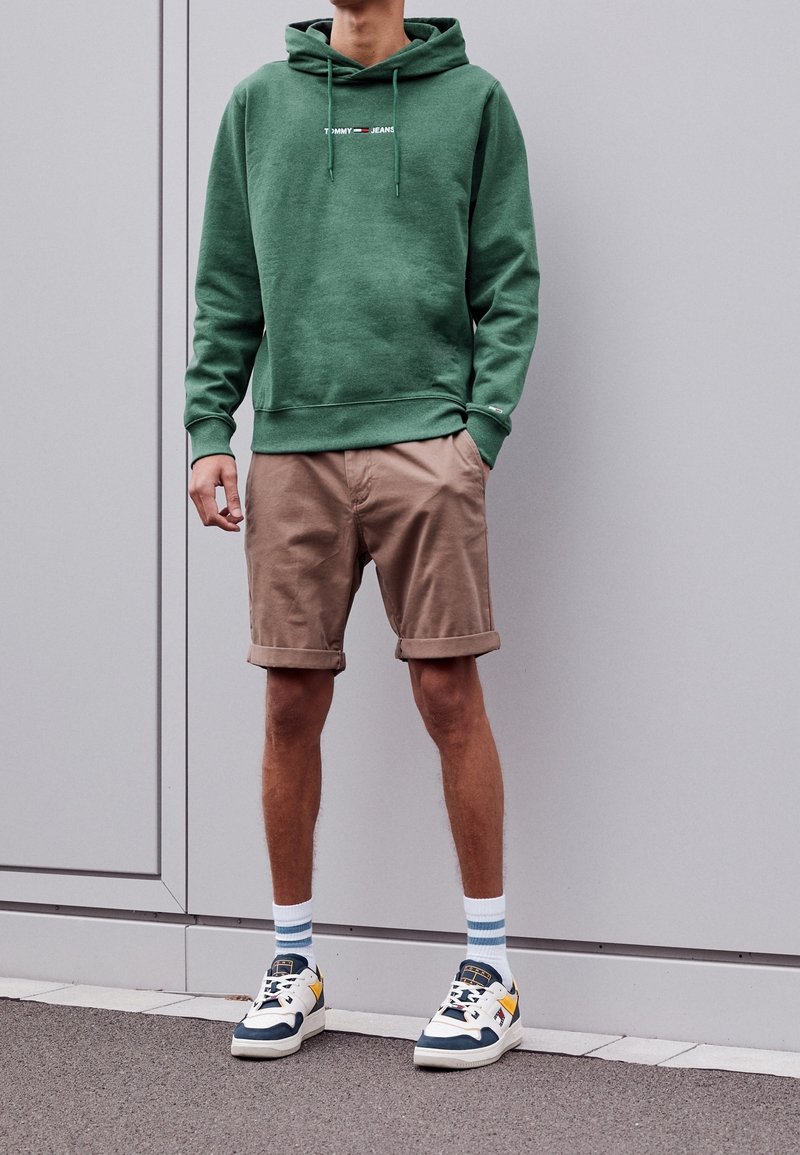 Grön hoodie med dragsko, beige uppvikta shorts och flerfärgade sneakers. Vita och blå randiga strumpor kompletterar outfiten.