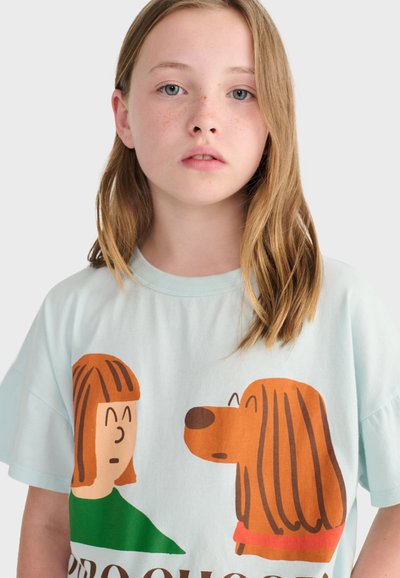 Młoda dziewczynka o jasnobrązowych włosach nosi jasnoniebieską koszulkę z kreskówkowymi ilustracjami przedstawiającymi osobę i psa zwróconych twarzami do siebie.