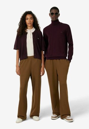 Zwei Models stehen nebeneinander und tragen braune Hosen mit weitem Bein; eines trägt ein weißes Poloshirt und eine dunkle Strickjacke, das andere einen dunklen Rollkragenpullover und eine Sonnenbrille.