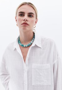 Camisa blanca de botones con una tela texturada, que presenta un bolsillo en el pecho. Accesorios incluyen un collar de cuentas en tonos azul y aqua y pendientes de aro dorados.