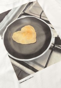 White T-shirt featuring a printed image of a heart-shaped pancake in a black frying pan, with a subtle kitchen background.