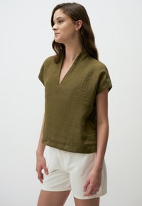 Top in lino verde oliva con scollatura a V e maniche corte, abbinato a pantaloni corti bianchi. Il tessuto presenta una finitura testurizzata e una forma rilassata.