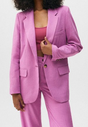 Vrouw draagt een roze pak met blazer en broek, gecombineerd met een donkerder roze cropped tanktop, tegen een effen achtergrond.