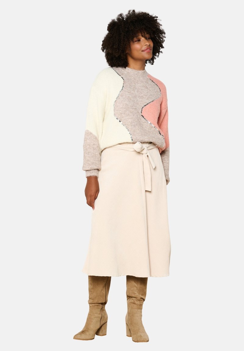 Mehrfarbiger Pullover mit einem wellenförmigen Design in Beige, Rosa und Creme, kombiniert mit einem cremefarbenen Rock und taupefarbenen kniehohen Stiefeln.