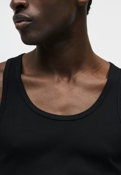 Κοντινό πλάνο ενός ατόμου που φοράει μαύρο ριπ τοπ χωρίς μανίκια, που δείχνει το ντεκολτέ, τις κλείδες και το κάτω μέρος του προσώπου με φόντο μονόχρωμο.