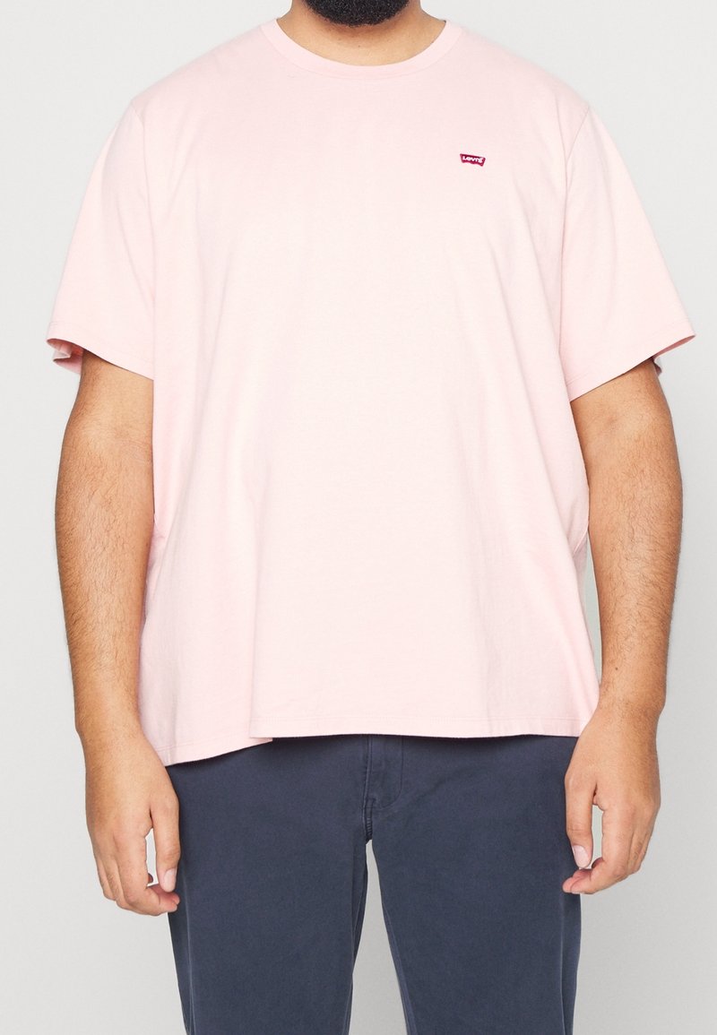 Homme portant un t-shirt Levi’s rose clair et un pantalon bleu foncé, debout les bras détendus le long du corps devant un fond uni.