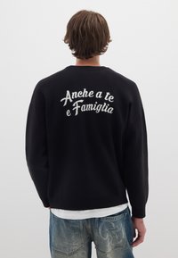 Maglione nero con maniche lunghe e colletto a giro. Sul retro è presente testo ricamato bianco "Anche a te e Famiglia." Texture morbida, vestibilità comoda.