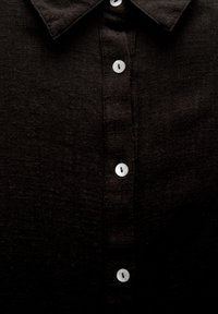Chemise noire en lin avec col classique, fermeture à boutons sur le devant avec des boutons blancs, et une surface de tissu texturée.