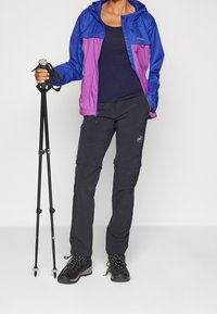 Colorful outdoor jacket in blue and pink, paired with black zippered pants and sturdy black hiking boots. Holding trekking poles.