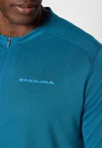 Teal cyklistický dres vyrobený z hladkej tkaniny, s polovičným zipsom vpredu, priedušnými sieťovinovými ramennými panelmi a logom "ENDURA" v svetlomodrej farbe.