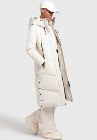 Manteau long en duvet crème matelassé avec capuche, fermetures éclair et boutons-pression, accents noirs et poches latérales. Porté avec un pantalon beige et des chaussures.