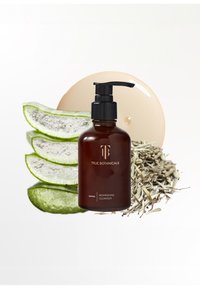 Bottiglia di ambra scura con pompa nera, etichettata "True Botanicals Nourishing Cleanser," accanto a fette di aloe fresche e elementi erboristici.