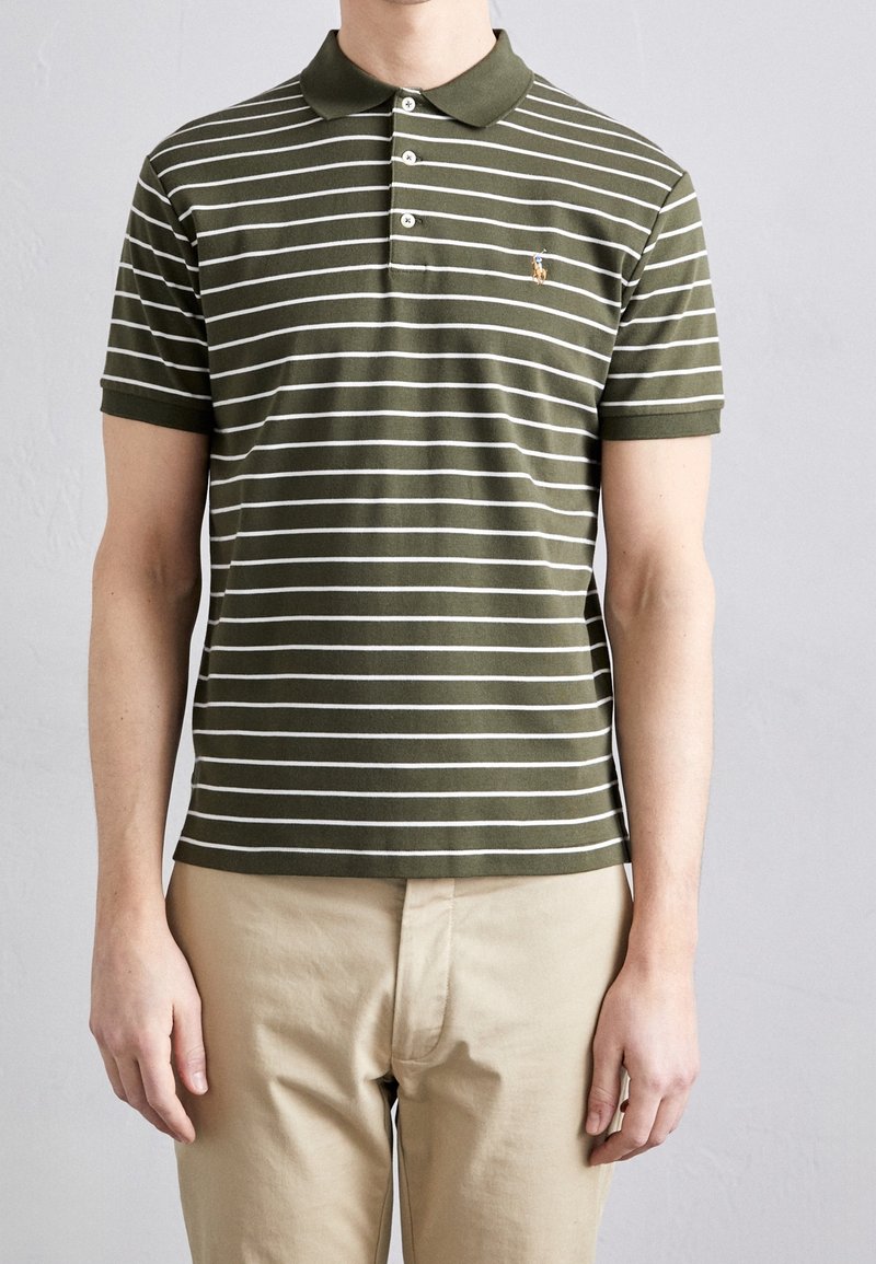 Homme portant un polo à manches courtes rayé vert olive et blanc avec un pantalon beige, debout contre un mur clair et uni.