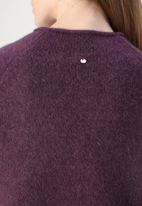 Violetti neulepaita, jossa on pörröinen tekstuuri, pyöreä pääntie ja pieni logokoriste niskassa.