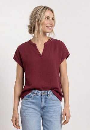 Mujer con cabello rubio recogido en un moño suelto, lleva una blusa burdeos de manga corta y vaqueros azul claro, sonriente y mirando hacia la derecha.