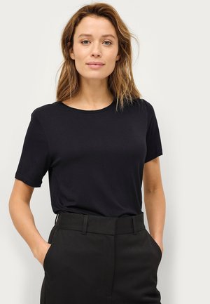 Woman with light brown hair wearing a plain black short-sleeve shirt and black high-waisted pants, standing with hands in pockets.